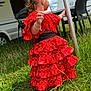 toddler, red_dress, flamenco_style, polka_dots, grass, outdoor, camper, child, young_child, back_view, fence_post, chair, black_chair, nature, summer, sunny, curly_hair, bracelet, dress, playing