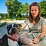 dog, woman, person, park, outdoor, tongue, harness, blanket, sitting, smile, pet, leash, trees, sunny_day, summer, portrait, happy, grass, friendship, casual_clothing