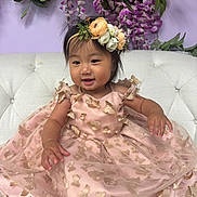 Eleanor joined the competition — help win amazing prizes! baby, child, dress, pink_dress, butterflies, flower_headband, floral_backdrop, couch, smiling, happy, portrait, indoor, cute, toddler, sitting, decor, greenery, purple_flowers, headwear, person