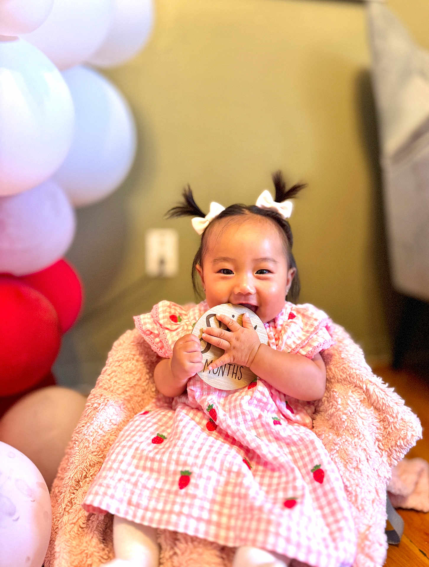 Eleanor is registered to the contest to win money with this photo: baby, child, girl, pigtails, bows, pink_dress, strawberry_pattern, blanket, balloons, indoor, happy, smiling, holding, wooden_sign, sitting, cute, portrait, infant, celebration, soft_texture