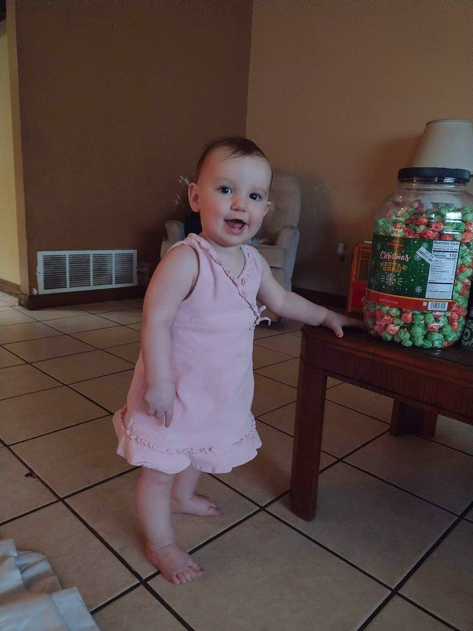 Sage is registered to the contest to win money with this photo: baby, baby_toddler_clothing, barefoot, child, floor, flooring, foot, hardwood, human_leg, kitchen_appliance, knee, pattern, person, pink, plastic_bottle, room, smile, standing, stool, toddler