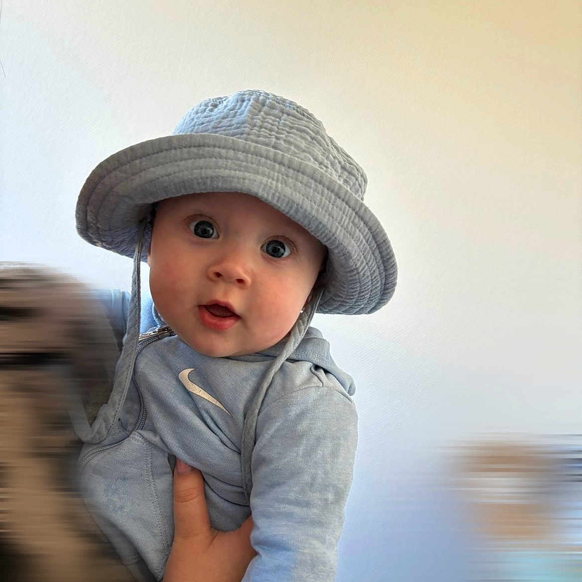 Lucyano a rejoint le concours — aidez-le/la à gagner de superbes lots ! adult_hand, baby, blue_clothing, bucket_hat, child, close_up, cute, hat, holding, hoodie, indoor, infant, nike_logo, photography, plain_wall, portrait, smile, soft_lighting, surprised_expression, wide_eyes
