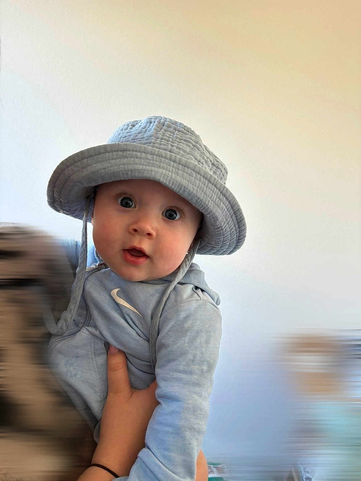 Lucyano a rejoint le concours — aidez-le/la à gagner de superbes lots ! baby, infant, child, bucket_hat, hat, blue_clothing, hoodie, nike_logo, wide_eyes, surprised_expression, holding, adult_hand, portrait, close_up, indoor, plain_wall, cute, smile, photography, soft_lighting