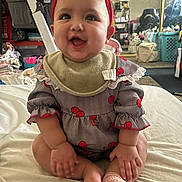 Alicia is registered to the contest to win money with this photo: baby, child, red_bow, bib, smiling, sitting, indoors, clothing, cute, happy, infant, face, hands, footwear, sock, bed, fabric, room, mirror, reflection