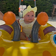 Ruby a rejoint le concours — aidez-le/la à gagner de superbes lots ! baby, yellow_headband, bow, inflatable_float, yellow, outdoor, greenery, child, cute, smile, play, summer, sunlight, person, infant, fun, water_toy, seat, happy, portrait