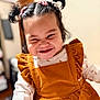 child, toddler, smiling, portrait, pigtails, hair_bows, corduroy, orange_dress, ruffles, cute, cheeks, indoor, close_up, happy, playful, eyes_squinting, long_sleeve_shirt, baby, background_blur, expression