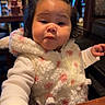 baby, background_people, black_hair, booth, child, close_up, cozy, cute, expression, face, fluffy_jacket, hands, indoor, menu, pink_bows, portrait, pout, restaurant, toddler, wooden_table
