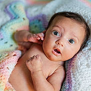 Mila is registered to the contest to win money with this photo: baby, infant, newborn, blanket, crochet_blanket, pastel_colors, surprised_expression, wide_eyes, portrait, closeup, skin, hand, lying_down, cute, cozy, soft_texture, nursery, bokeh, photography, face