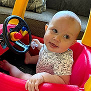 Honey joined the competition — help win amazing prizes! baby, child, toy_car, steering_wheel, pacifier, indoor, couch, cushion, red, yellow, playful, smiling, clothing, short_sleeve, person, infant, seat, hand, foot, portrait