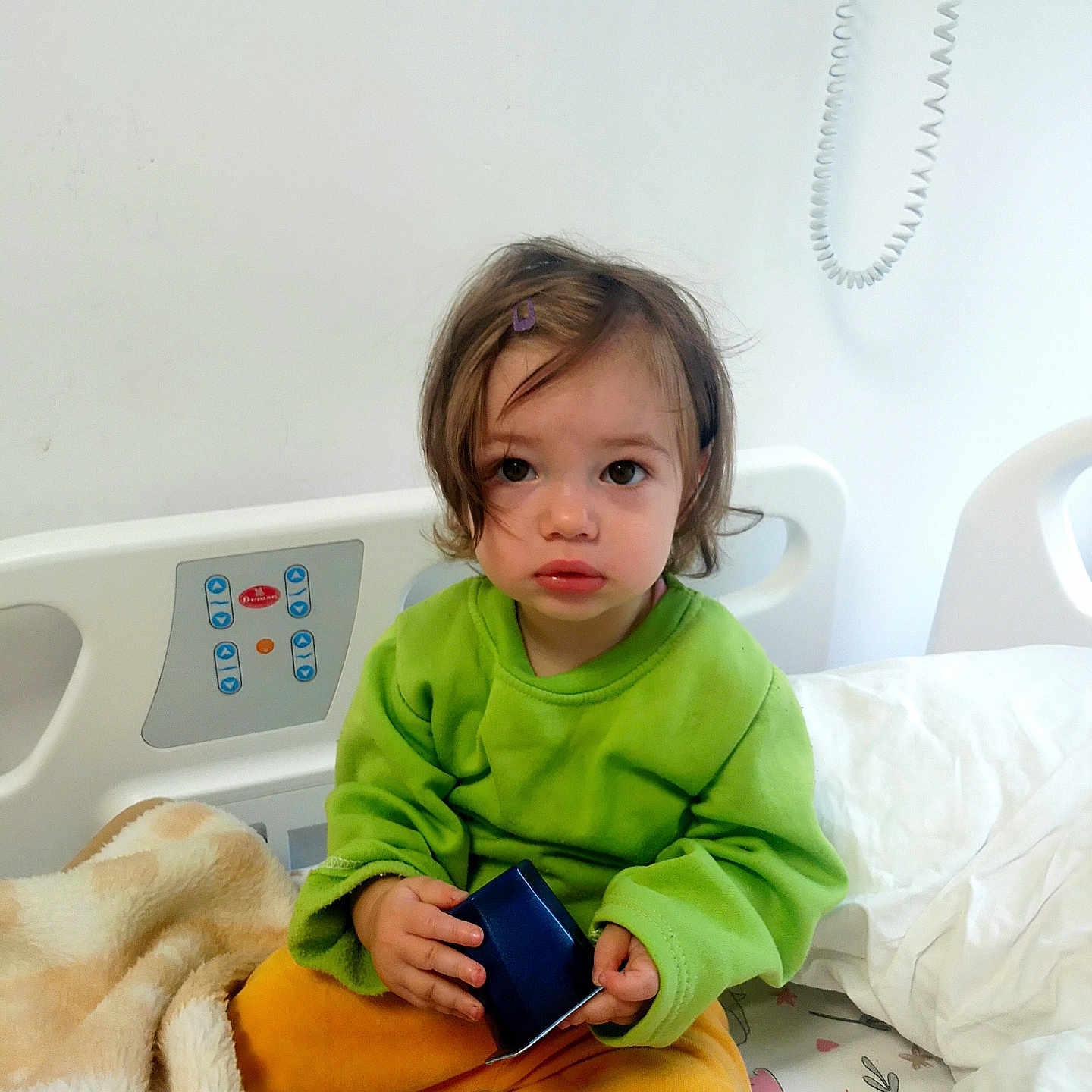 Artenisa participe au concours pour gagner de l'argent avec cette photo : bed, blanket, child, comfort, curly_hair, expression, face, green_sweater, hospital_bed, indoor, looking_at_camera, medical_bed, person, plush_blanket, sitting, toddler, toy, white_wall, yellow_pants, young_child