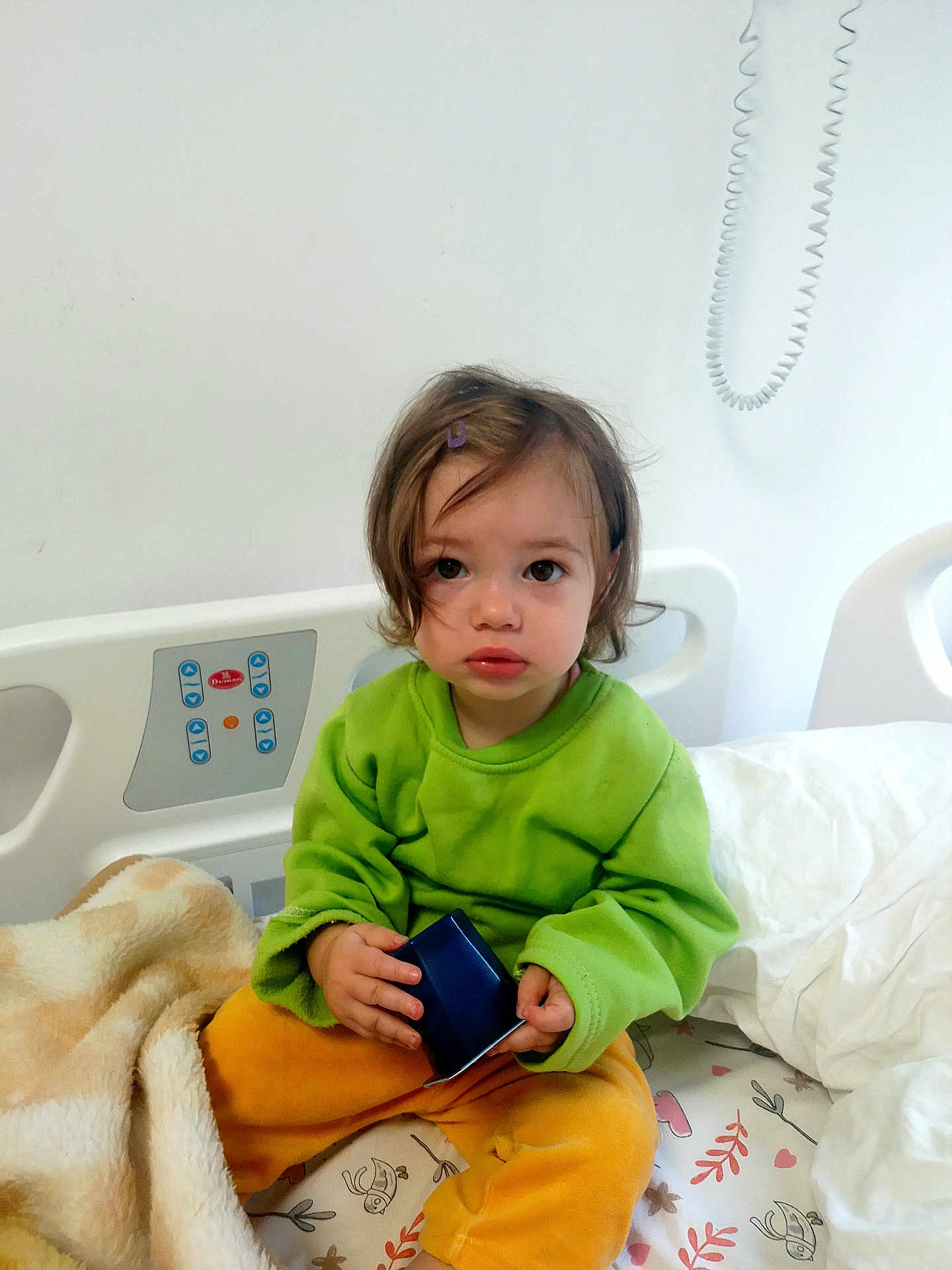 Artenisa participe au concours pour gagner de l'argent avec cette photo : toddler, child, hospital_bed, blanket, green_sweater, yellow_pants, toy, indoor, person, bed, curly_hair, face, looking_at_camera, young_child, comfort, plush_blanket, white_wall, medical_bed, expression, sitting