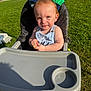 toddler, high_chair, outdoor, grass, sunlight, child, snack, seat, parked_cars, red_hair, baby_clothing, summer, playful, sunny_day, person, food, hand, expression, greenery, daylight
