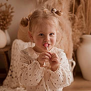 Lou participe au concours pour gagner de l'argent avec cette photo : child, girl, lollipop, blouse, hair_buns, smile, face, portrait, indoor, vase, decor, dried_plants, cozy, warm_tones, cute, hands, young, happy, soft_light, background_blur