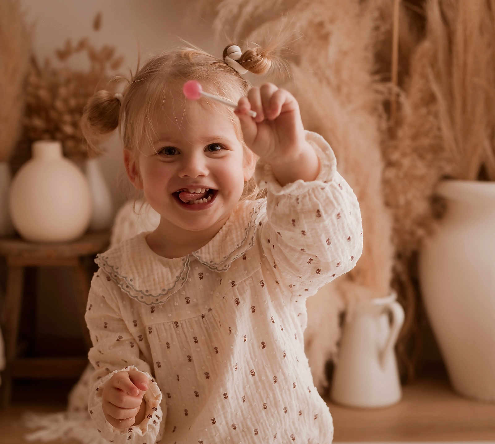 Lou participe au concours pour gagner de l'argent avec cette photo : toddler, child, smiling, lollipop, pigtails, indoor, home_decor, vase, dried_plants, cute, happy, portrait, warm_lighting, cozy, playful, white_clothing, patterned_dress, person, young_child, fun