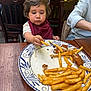 toddler, child, french_fries, plate, food, bib, table, wooden_chair, restaurant, yellow_wall, person, eating, meal, curly_hair, casual_clothing, hand, indoor, dining, appetite, focus