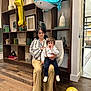 Attila is registered to the contest to win money with this photo: woman, child, toddler, balloon, chair, bookshelf, wood_floor, celebration, indoor, decor, number_one_balloon, shark_balloon, suspenders, white_shoes, striped_shirt, pants, person, home, seated, floor