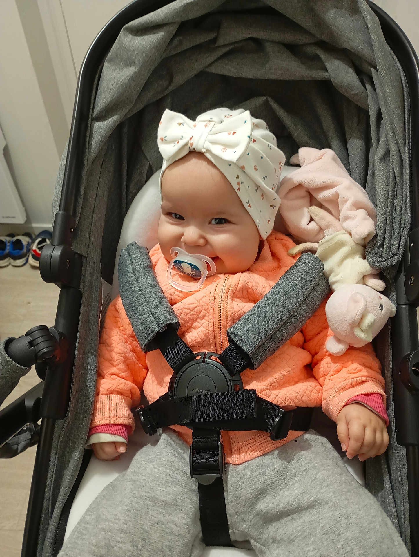 Elyana a rejoint le concours — aidez-le/la à gagner de superbes lots ! baby, pacifier, stroller, headwear, orange_jacket, gray_pants, smile, plush_toy, indoor, child, seatbelt, baby_bow, clothing, face, person, floor, shoes, hand, cute, infant
