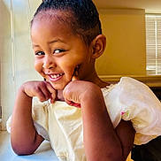Ogeti is registered to the contest to win money with this photo: child, girl, smiling, portrait, indoors, white_dress, curly_hair, hair_bun, hair_clips, hands_under_chin, happy, laughing, sitting, window, blinds, warm_lighting, close_up, fashion, photography, home