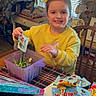 child, girl, yellow_sweatshirt, smile, candy, table, chair, window, blinds, container, packet, indoor, home, decorations, wooden_table, paper_towel, color_activity_set, happy, person, toy