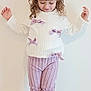 child, girl, white_sweater, lilac_bows, curly_hair, hair_clip, pink_pants, striped_pants, black_boots, standing, looking_down, shy_smile, indoor, cream_wall, carpet, full_body, portrait, kids_fashion, ribbon, cute
