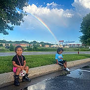 Jianna is registered to the contest to win money with this photo: child, kids, curb, puddle, rainbow, grass, tree, clouds, sky, reflection, boots, shorts, tshirt, sidewalk, street, parking_lot, urban, outdoor, smile, playful