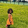 Jianna joined the competition — help win amazing prizes! toddler, child, orange_dress, grass, outdoor, sunlight, curly_hair, fence, shadow, greenery, building, side_view, young_child, summer, standing, casual_clothing, park, daylight, nature, person