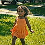 child, girl, orange_dress, grass, outdoor, sunlight, curly_hair, walking, sidewalk, tree, car, parked_car, summer, earrings, necklace, footwear, nature, person, young_child, daytime