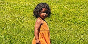 Jianna is registered to the contest to win money with this photo: child, grass, outdoor, dress, red_shoes, curly_hair, sunlight, fence, parked_cars, playing, summer, greenery, person, field, casual_clothing, young_child, daylight, standing, nature, urban_background