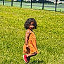 child, grass, outdoor, dress, red_shoes, curly_hair, sunlight, fence, parked_cars, playing, summer, greenery, person, field, casual_clothing, young_child, daylight, standing, nature, urban_background