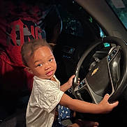 Alijah is registered to the contest to win money with this photo: child, toddler, car, steering_wheel, driver_seat, vehicle_interior, chevrolet, sitting, portrait, face, short_hair, white_shirt, blue_shorts, barefoot, hands, dashboard, night, low_light, indoor, playful