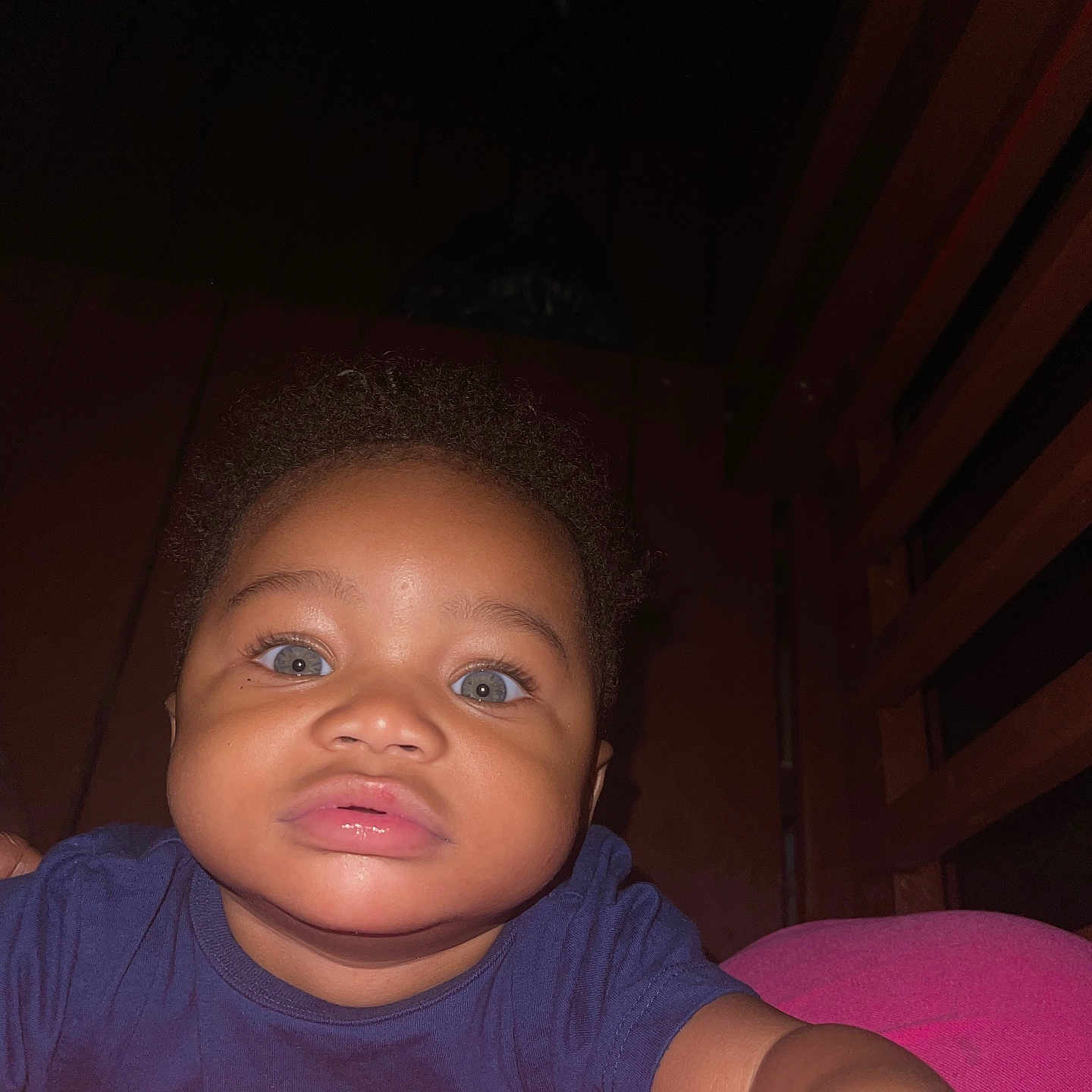 Alijah is registered to the contest to win money with this photo: baby, child, portrait, close_up, face, wide_eyed, serious_expression, indoor, dim_lighting, flash_photography, pink_cushion, blue_shirt, wooden_background, sitting, looking_at_camera, infant, dark_background, arms, headshot, casual_photo