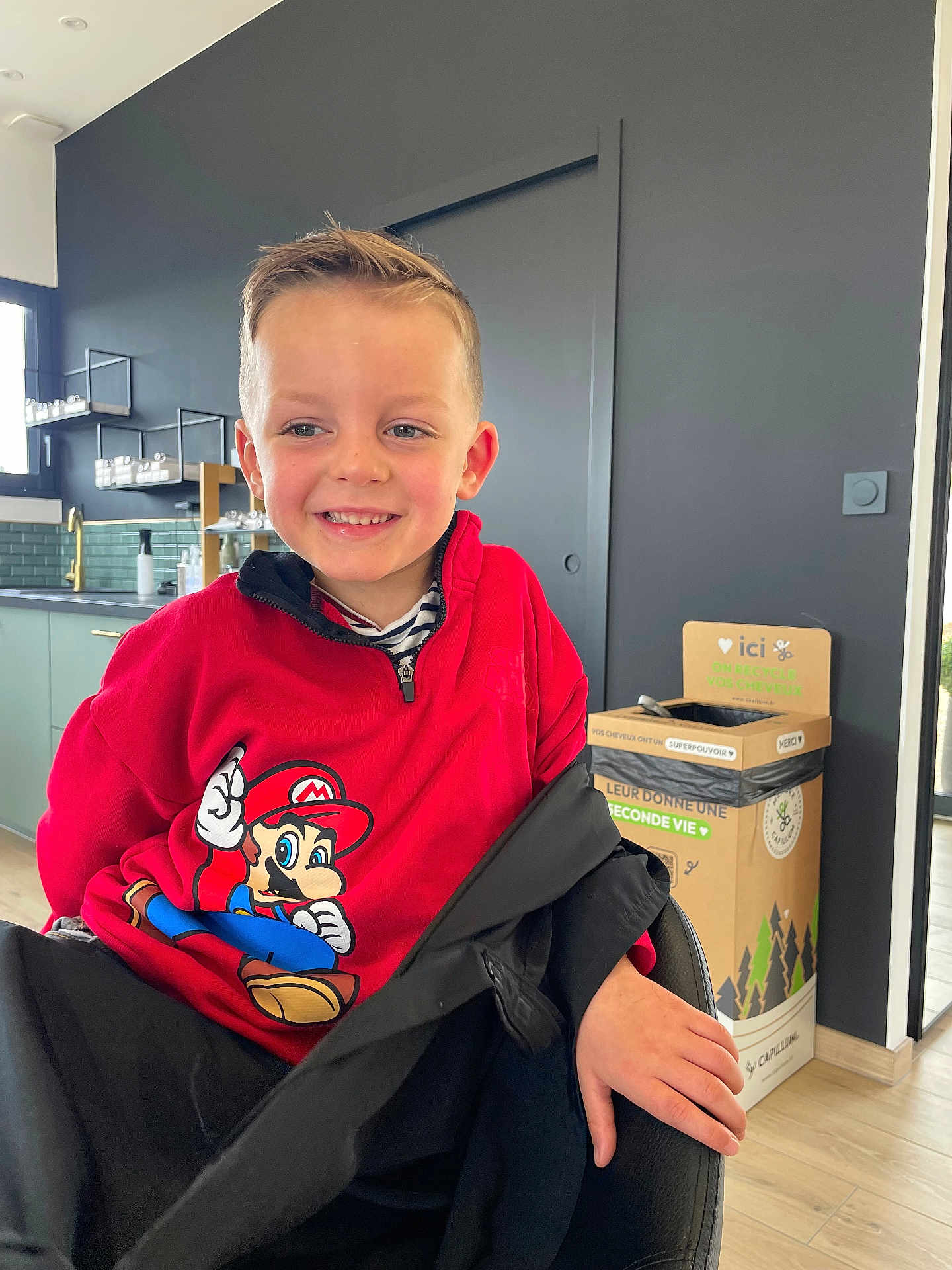 Matheo participe au concours pour gagner de l'argent avec cette photo : child, boy, smile, red_sweatshirt, super_mario, indoor, kitchen, modern, recycling_bin, black_jacket, happy, casual, seated, short_hair, person, light_skin, furniture, window, wood_floor, face