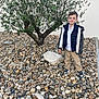 child, boy, tree, rocks, pebbles, outdoor, smiling, casual_clothing, jacket, sneakers, nature, daylight, wall, ground, portrait, standing, young, greenery, leaves, background