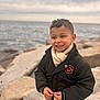 child, smiling_child, curly_hair, scarf, jacket, coat, hands, rocks, sea, ocean, horizon, overcast, cloudy_sky, portrait, outdoor, waterfront, smile, cold_weather, bokeh, coastline