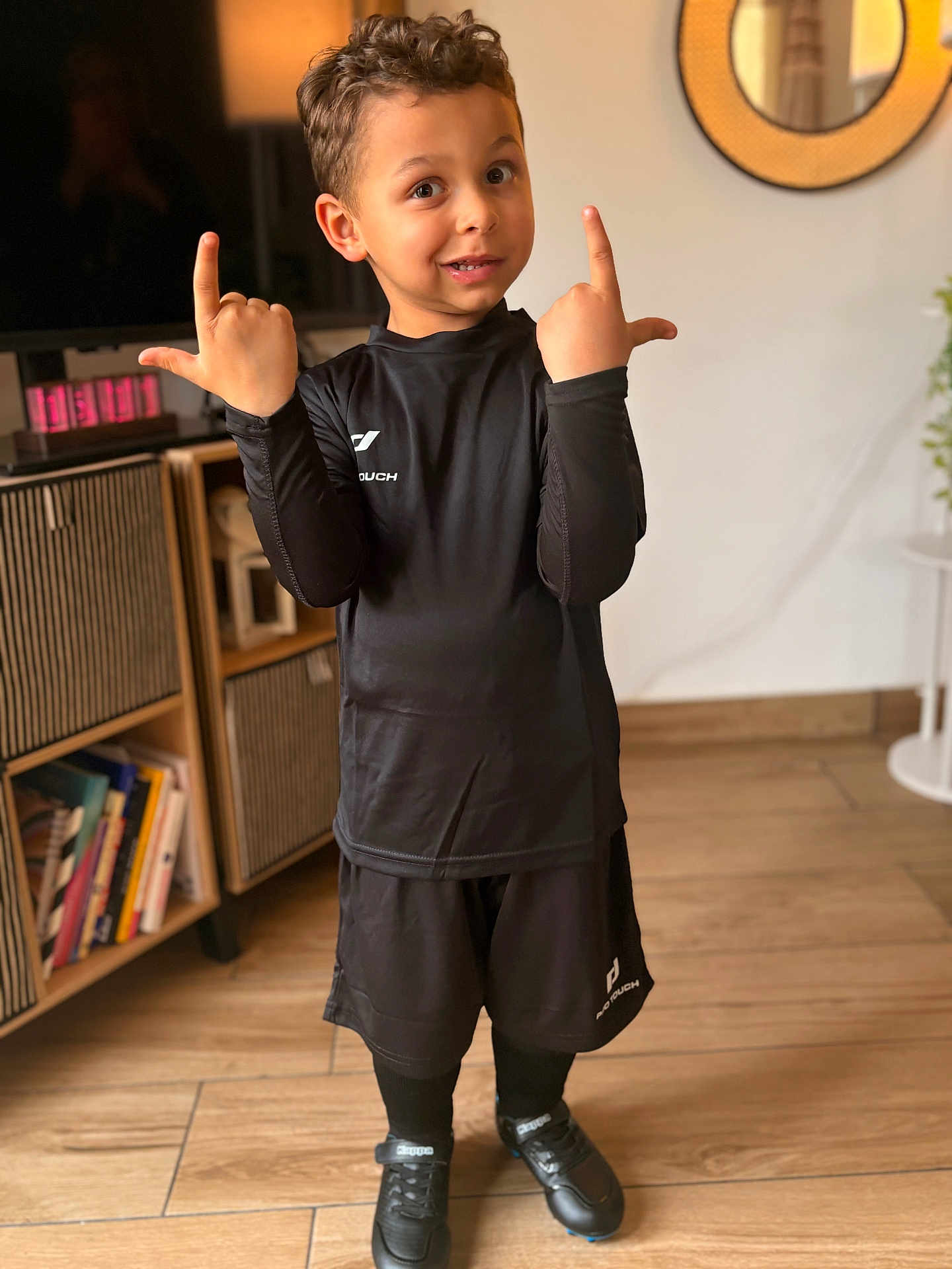 Tom participe au concours pour gagner de l'argent avec cette photo : child, boy, smile, portrait, indoor, black_uniform, soccer_cleats, shorts, long_sleeve, socks, hands_up, curly_hair, brown_eyes, living_room, bookshelf, mirror, cabinet, wooden_floor, playful, cute