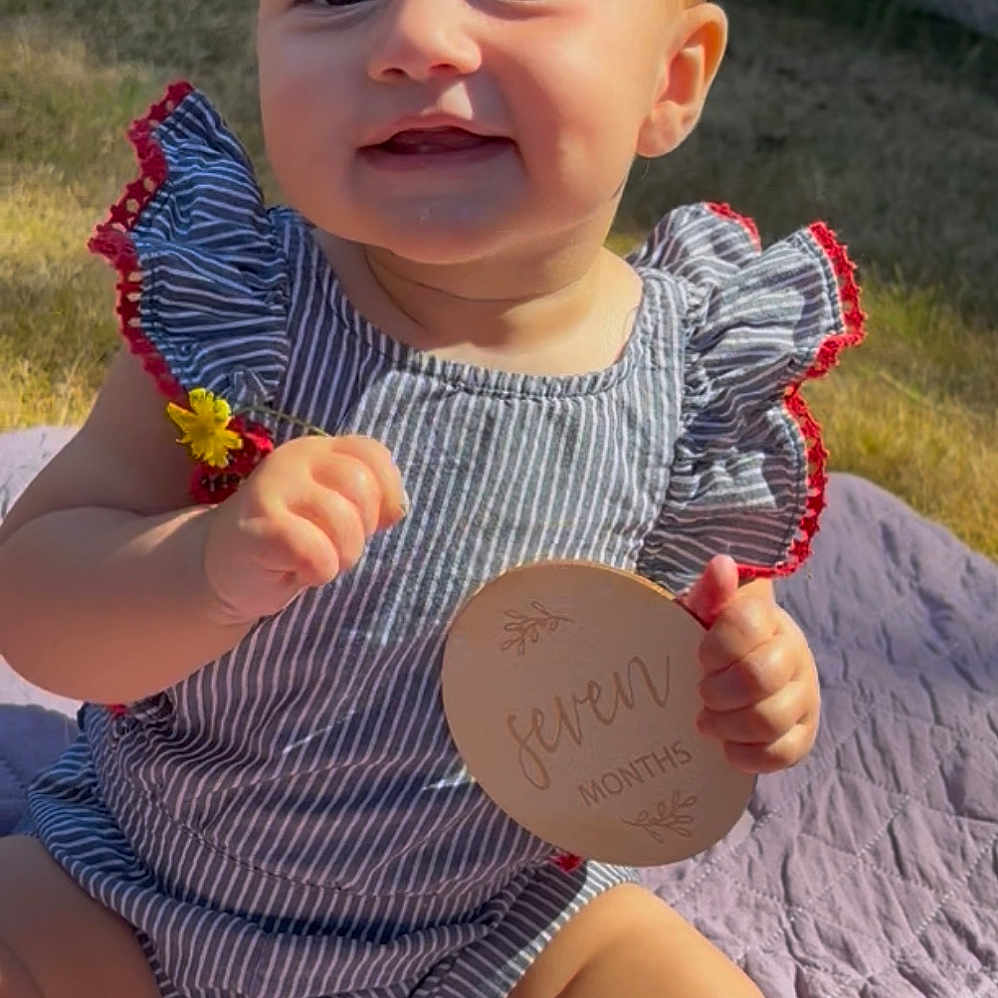 Analeigh joined the competition — help win amazing prizes! baby, child, cute, fence, flower, grass, hands, happy, infant, nature, outdoor, person, portrait, quilt, sitting, smiling, striped_clothing, summer, sunlight, wooden_sign