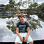 Kelyan participe au concours pour gagner de l'argent avec cette photo : boy, child, smile, pond, tree, water, reflection, cloudy_sky, casual_clothing, shorts, tshirt, outdoor, nature, urban, bench, sitting, relaxed, daytime, background_blur, young