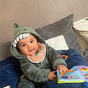 Issa a rejoint le concours — aidez-le/la à gagner de superbes lots ! baby, child, dinosaur_onesie, hood, plush_teeth, bib, soft_book, couch, cushion, indoor, smiling, happy, playing, toy, infant, person, clothing, fabric, cute, portrait