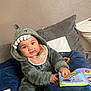 baby, child, dinosaur_onesie, hood, plush_teeth, bib, soft_book, couch, cushion, indoor, smiling, happy, playing, toy, infant, person, clothing, fabric, cute, portrait