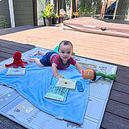 Zachary is registered to the contest to win money with this photo: baby, child, play_mat, toy, book, outdoor, deck, sunlight, smiling, face, clothing, plant, remote_control, blanket, wooden_floor, potted_plant, red_toy, blue_shorts, striped_shirt, curious
