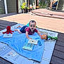 baby, child, play_mat, toy, book, outdoor, deck, sunlight, smiling, face, clothing, plant, remote_control, blanket, wooden_floor, potted_plant, red_toy, blue_shorts, striped_shirt, curious