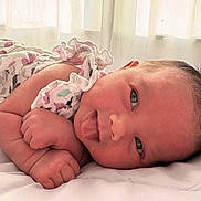 Josee is registered to the contest to win money with this photo: baby, infant, face, tongue_out, skin, hair, clothing, floral_pattern, ruffles, lying_down, bed, blanket, soft_light, curtains, indoors, cute, expression, newborn, portrait, sleepy