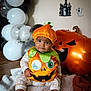 baby, child, pumpkin_costume, halloween, balloons, inflatable, jack_o_lantern, ghost_decorations, blanket, floor, costume, holiday, indoor, cute, sitting, expression, orange, green, black, white