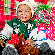 Kei'Jon joined the competition — help win amazing prizes! baby, child, toddler, smiling, green_jacket, beanie, plush_toy, reindeer, christmas, holiday, decorations, red_bow, black_rug, sitting, happy, indoor, festive, cute, person, portrait