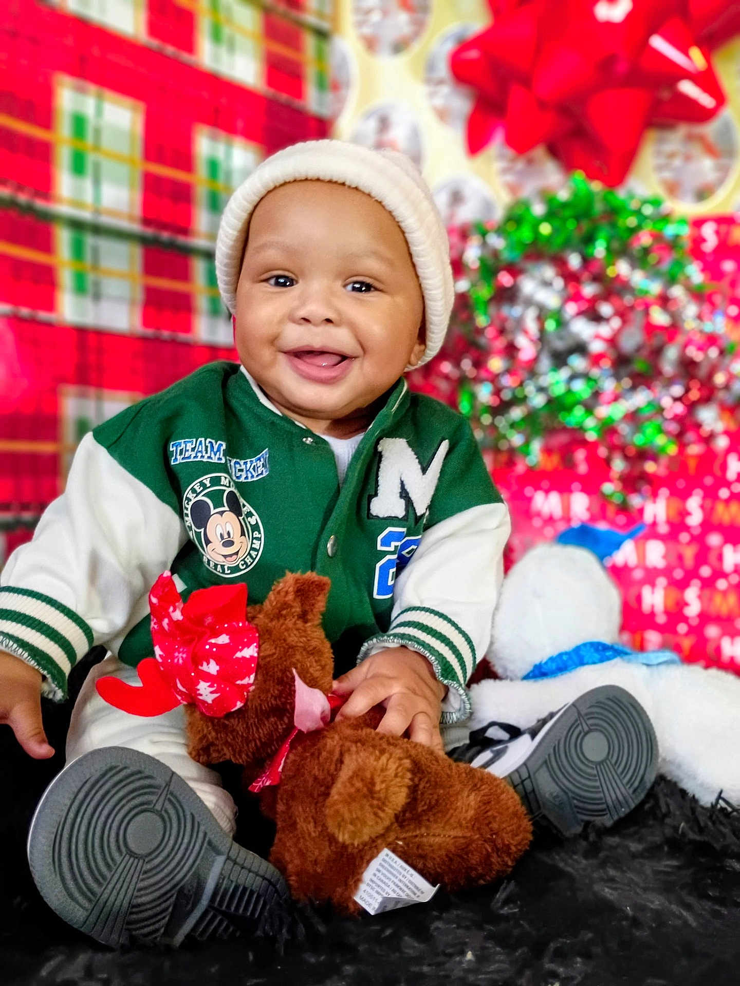 Kei'Jon joined the competition — help win amazing prizes! baby, child, toddler, smiling, green_jacket, beanie, plush_toy, reindeer, christmas, holiday, decorations, red_bow, black_rug, sitting, happy, indoor, festive, cute, person, portrait