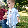 child, toddler, outdoor, grass, tree, person, adult, shirt, pants, sneakers, sunlight, shadow, nature, park, casual_clothing, greenery, daylight, curious, standing, relaxing