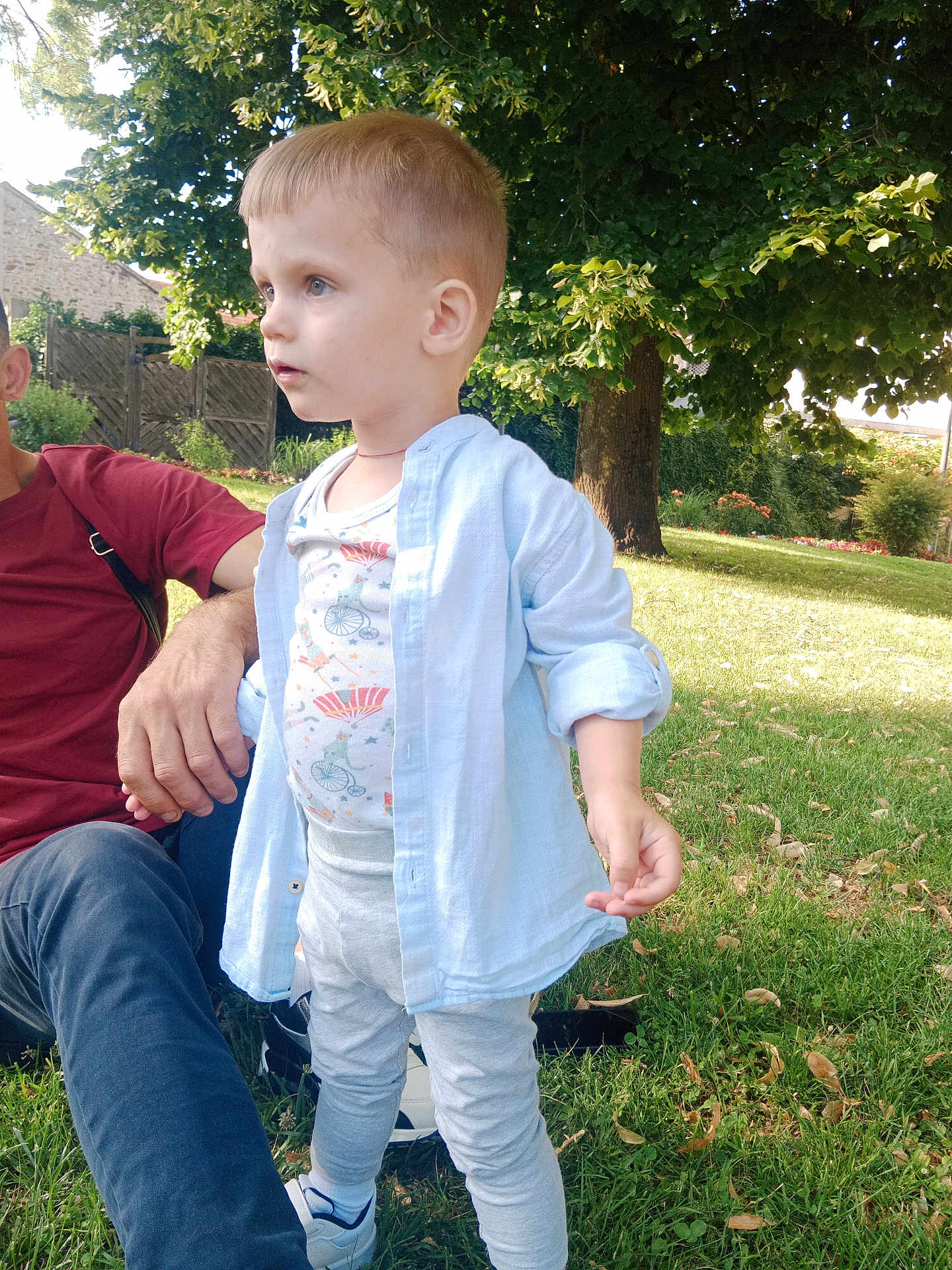 Teo participe au concours pour gagner de l'argent avec cette photo : child, toddler, outdoor, grass, tree, person, adult, shirt, pants, sneakers, sunlight, shadow, nature, park, casual_clothing, greenery, daylight, curious, standing, relaxing