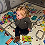 balls, blonde_hair, book, brown_shoes, casual_clothing, child, colorful, curious, curly_hair, cute, exploration, floor, fun, indoor, playing, playmat, sitting, toddler, toys, young_child