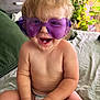 baby, toddler, child, purple_sunglasses, heart_shaped_glasses, smiling, happy, curly_hair, diaper, sitting, bed, blanket, green_pillow, plant, indoor, cute, playful, hands, face, skin