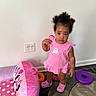 carpet, chair, child, crocs, curly_hair, cute, expression, floor, home, indoor, minnie_mouse, orange_ring, person, pink_dress, play_area, purple_ring, standing, toddler, toy, wall_outlet